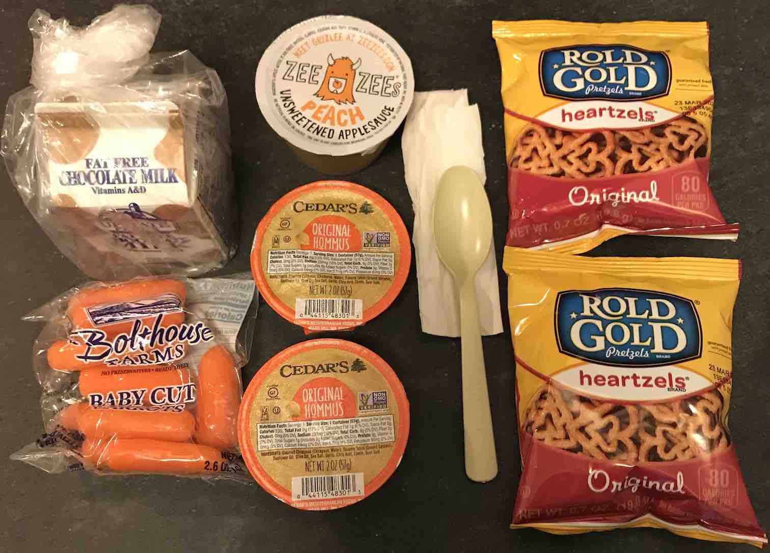 meal containing hummus, baby carrots, 2 pouches of pretzels, chocolate milk, apple and peach sauce, a paper napkin, and a plastic spoon